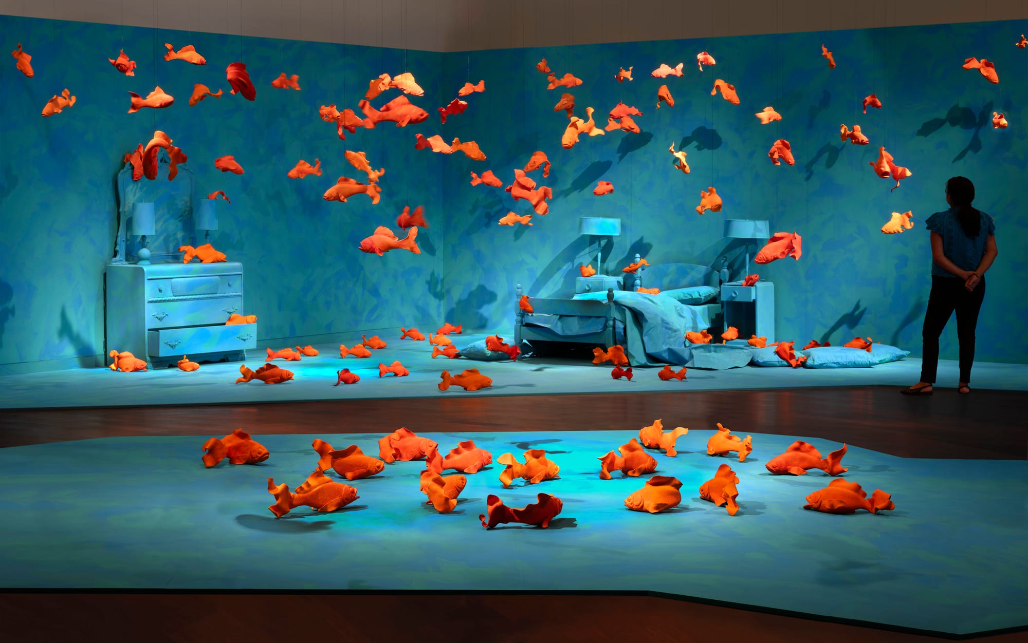 Installation view of Sandy Skoglund’s Revenge of the Goldfish at the McNay Art Museum, featuring bright orange ceramic fish suspended in a blue bedroom scene.