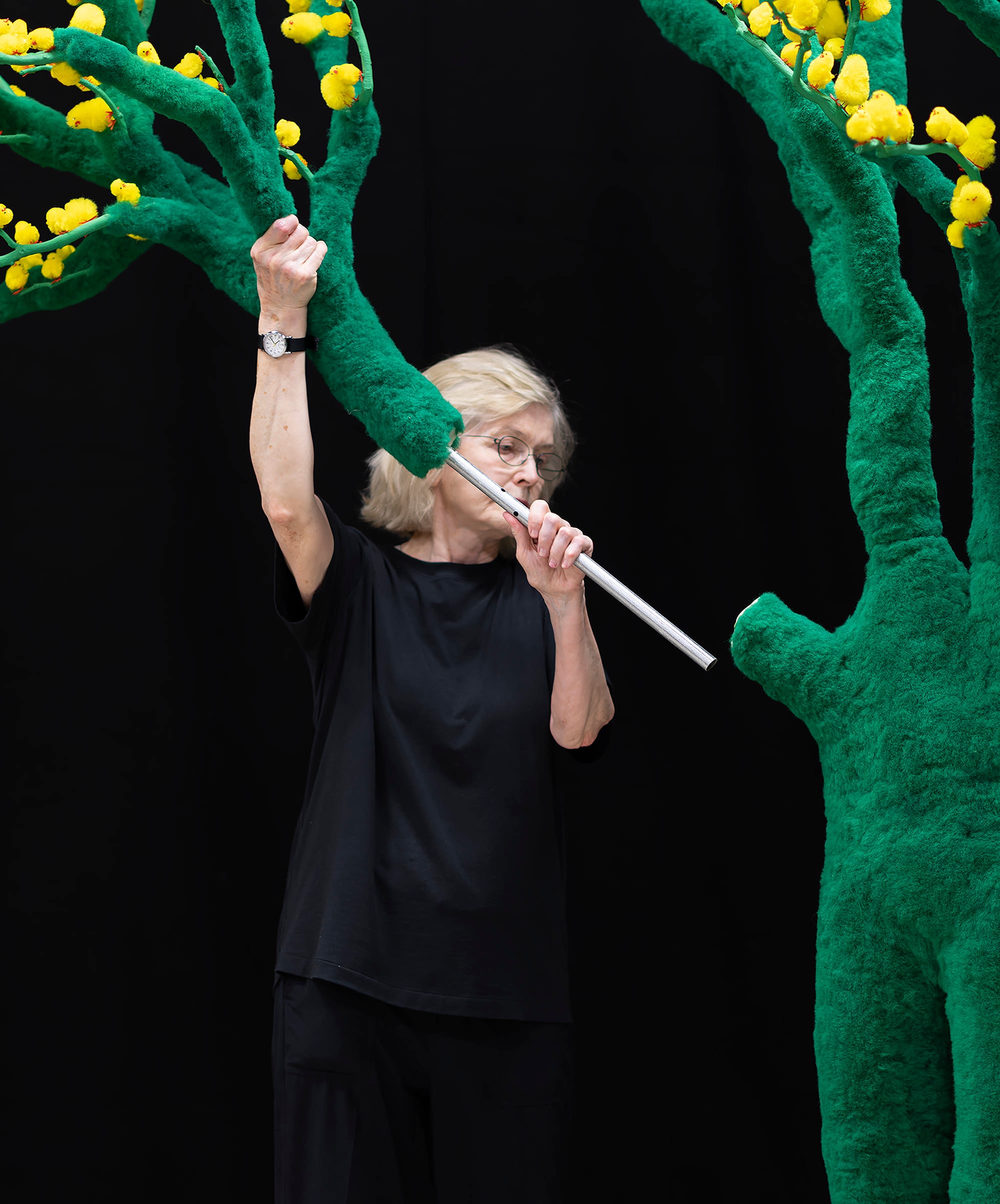 An woman wearing black assembles a green sculptural tree with yellow blossoms, inserting a metal branch into its trunk against a dark background.