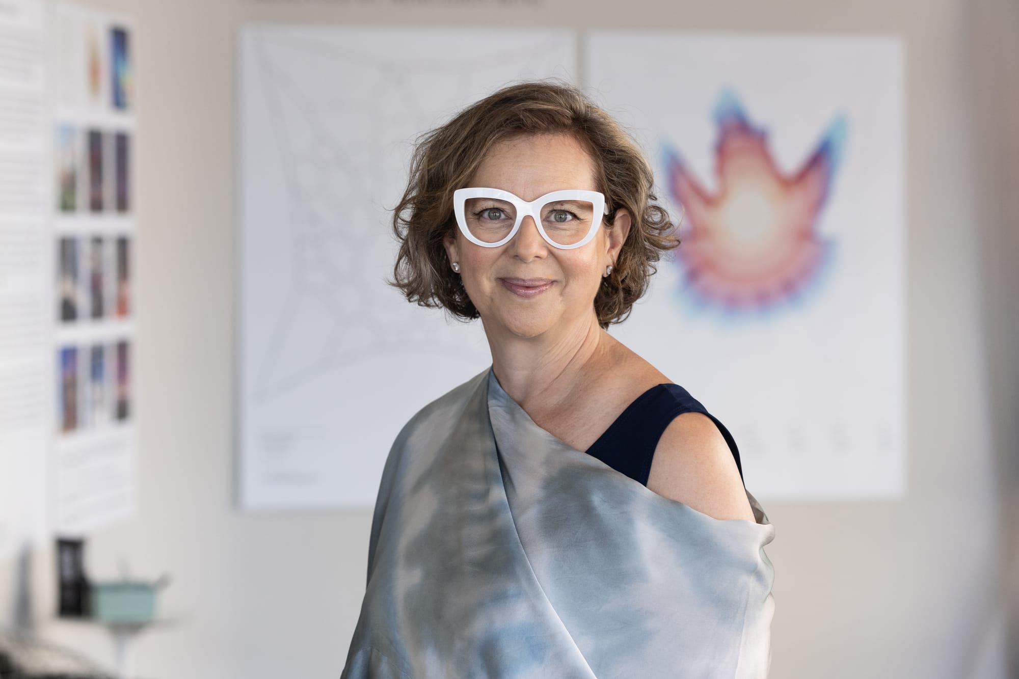 Portrait of artist Janet Echelman wearing white glasses and a draped gray shawl, standing in her studio with artwork diagrams in the background.