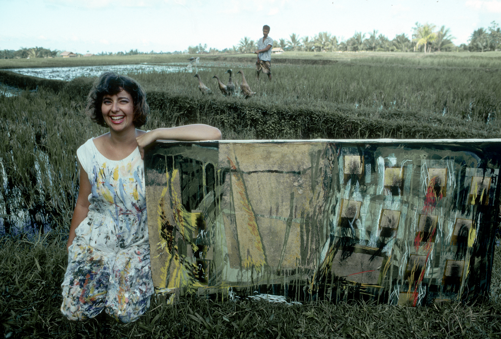 Janet Echelman smiling in a paint-splattered dress beside one of her early abstract paintings in a lush rice field in Bali, with a farmer and ducks in the background.