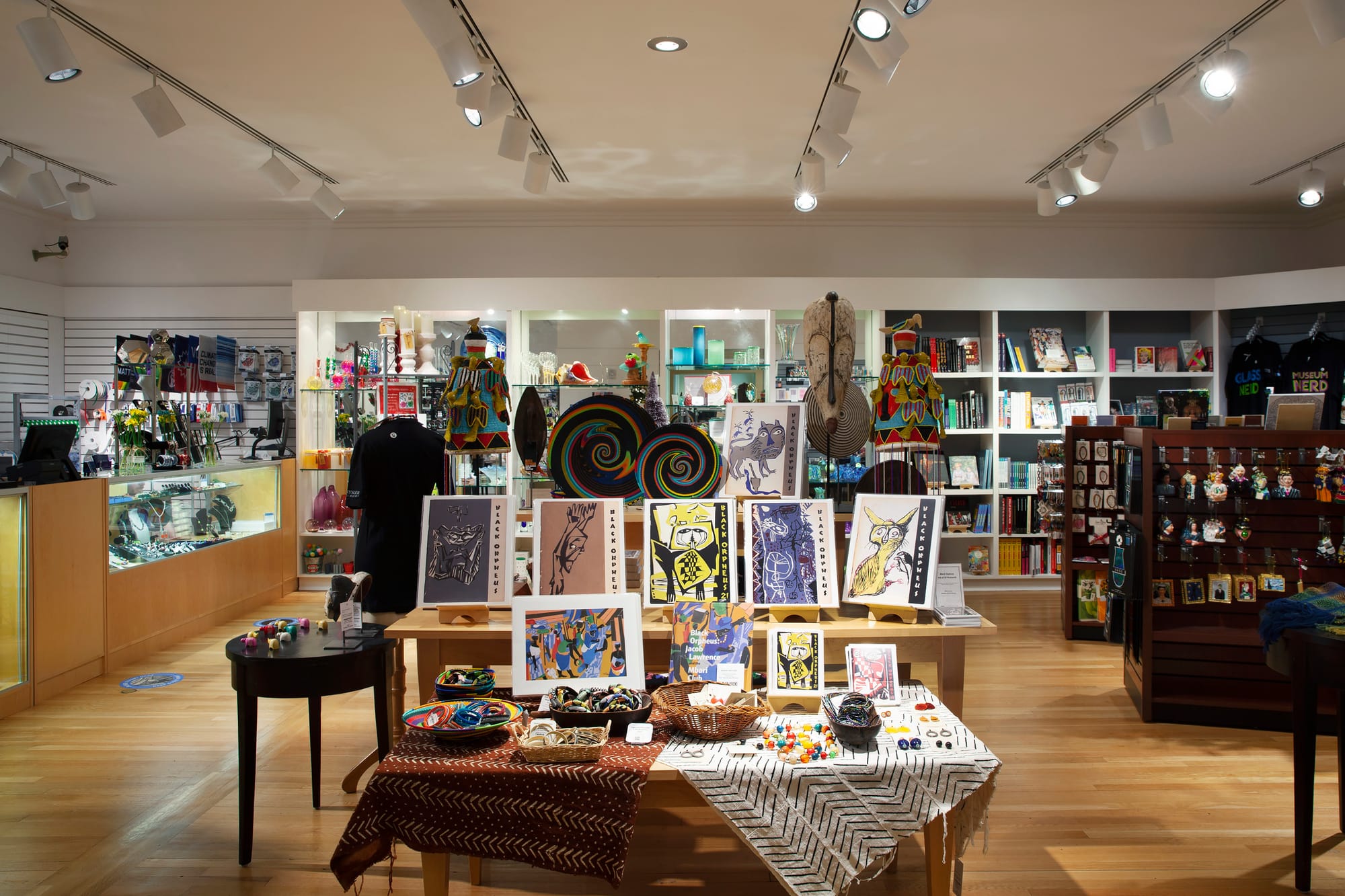 Interior of a well-lit museum gift shop with tables displaying colorful prints, crafts, and jewelry, surrounded by shelves of books, art objects, and merchandise.