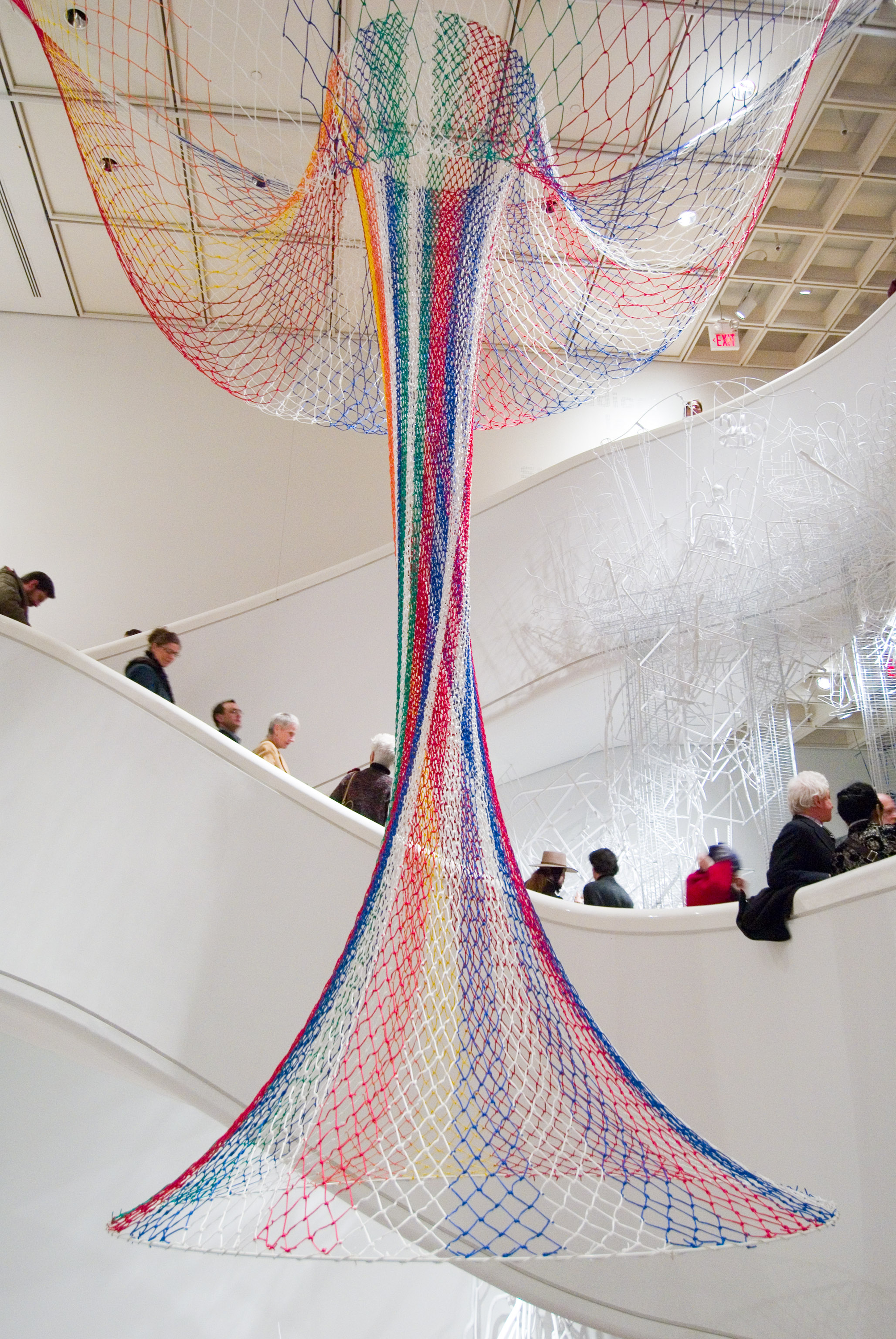 Janet Echelman’s suspended net sculpture made of colorful woven cords stretching between two levels of a museum atrium as visitors walk along a curved ramp below.