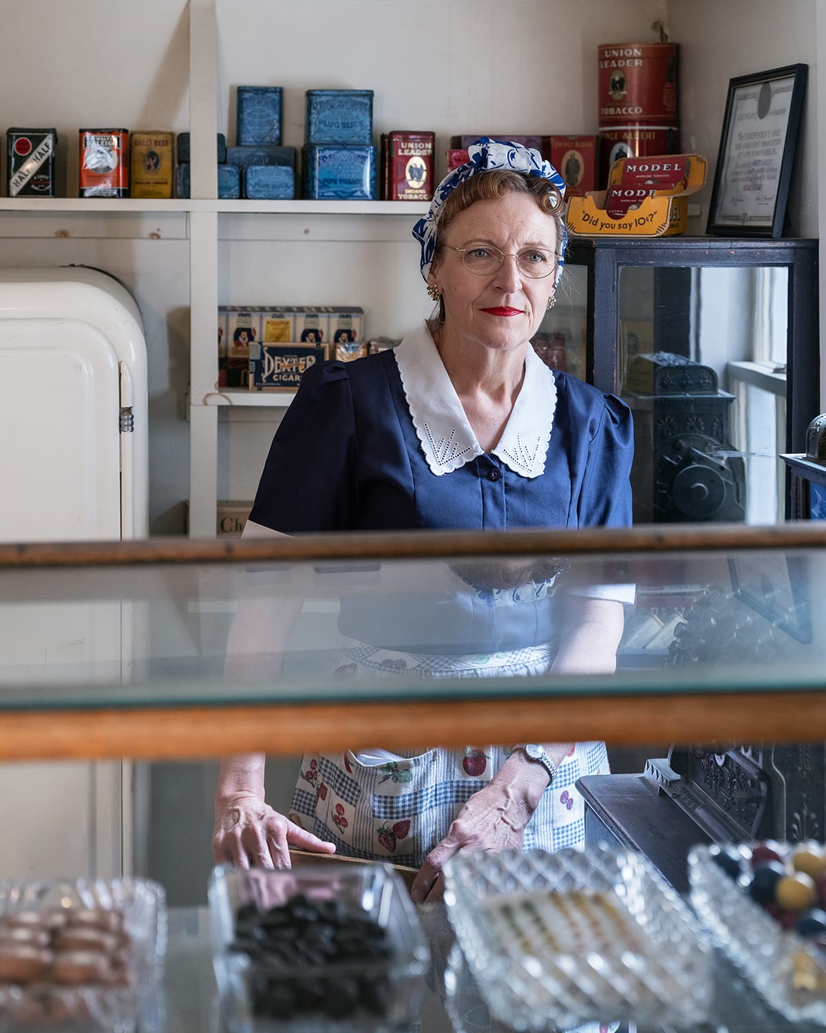 A woman dressed in 1940s-style clothing stands behind a glass candy counter in a vintage shop filled with old tobacco tins and memorabilia.