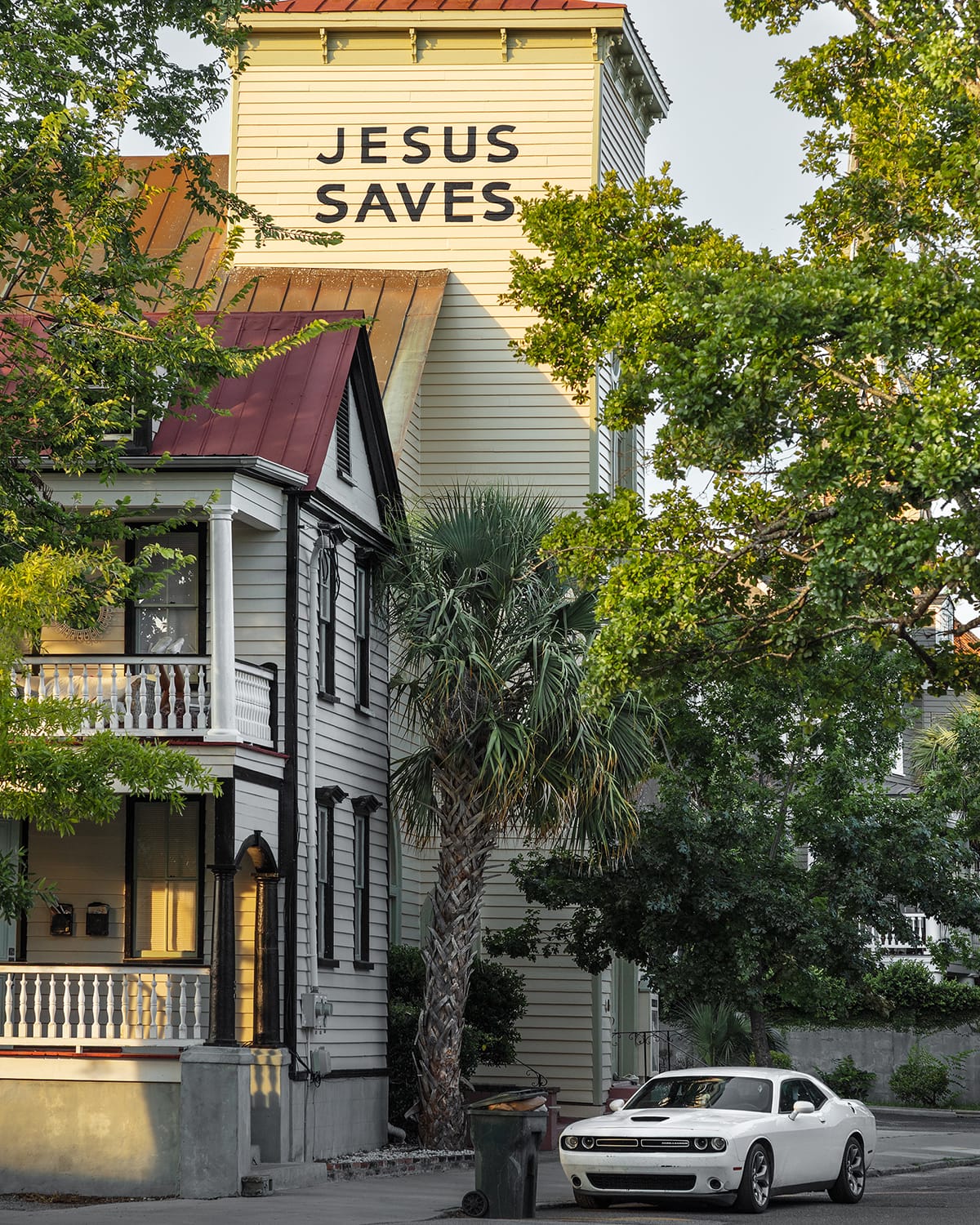 A pale yellow church tower with the words “JESUS SAVES” rises above houses and palm trees, with a white car parked below.