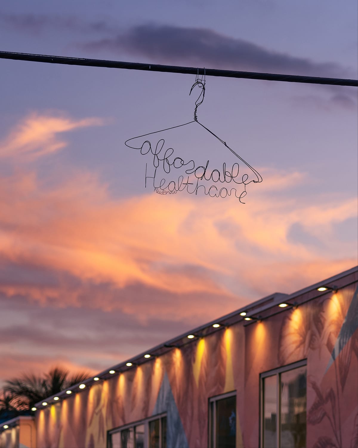 A wire hanger shaped to spell “affordable healthcare” hangs against a colorful sunset sky above a mural-lit building.
