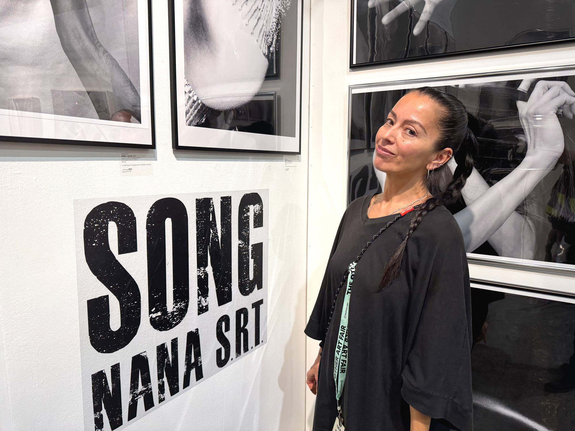 A woman stands in an exhibition space smiling and her head tilted in front of the text "SONG NANA S.R.T."