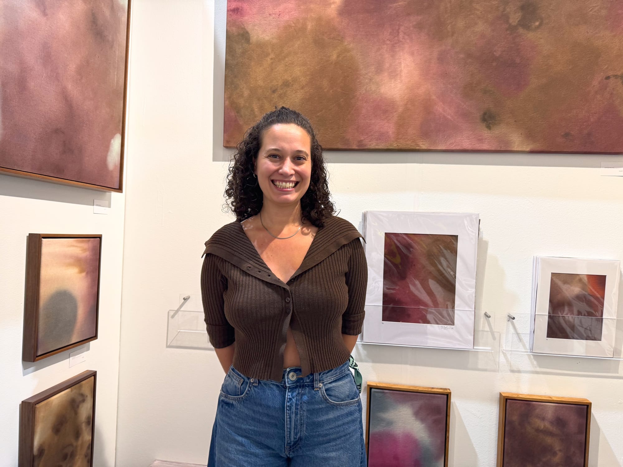 A woman stands with her arms behind her back smiling in front of her abstract artworks in a gallery setting