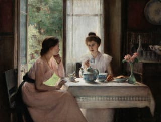Two women sit at a sunlit table having tea beside an open doorway, with one woman leaning on her hand and listening as the other smiles and holds a teacup.