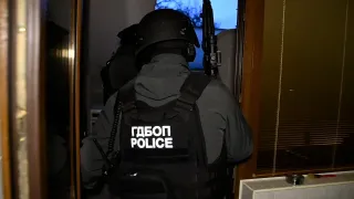 A police officer in tactical gear and a helmet enters a dimly lit building through a doorway, carrying a long firearm, with Cyrillic lettering and the word “POLICE” on the back of his vest.