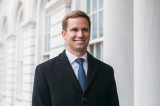 A man with a short haircut, a black suit and blue tie smiles in front of a government building.