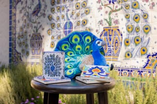 Plush peacock, peacock-patterned mug, and decorative tile displayed on a stool in front of a colorful peacock-themed mosaic wall.