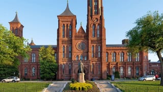 Red-brick Smithsonian Castle on the National Mall, framed by green trees, with a statue at the front entrance under a clear blue sky.