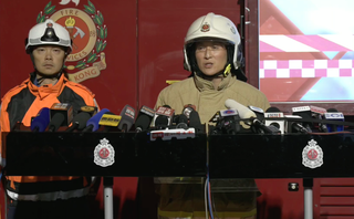 wo Hong Kong Fire Services officers stand at a podium during a nighttime press briefing, surrounded by microphones, with a fire services emblem and emergency vehicle visible behind them.
