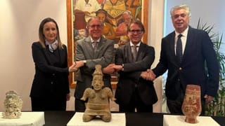 Four officials shake hands behind three pre-Hispanic artifacts displayed on tables during a restitution ceremony at the Mexican Embassy in Lisbon