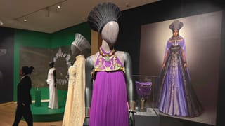 Mannequin dressed in a regal purple Wakanda gown and ornate headpiece on display in a museum gallery, with a large portrait of the character in the background and additional costumes nearby.