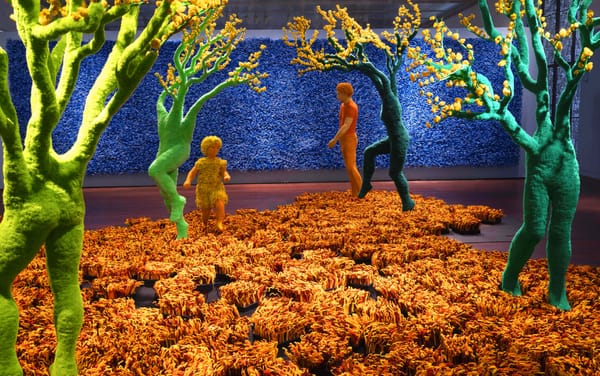 Colorful installation features life-size human figures and tree like forms. The floor is orange and the wall is a blue and textured
