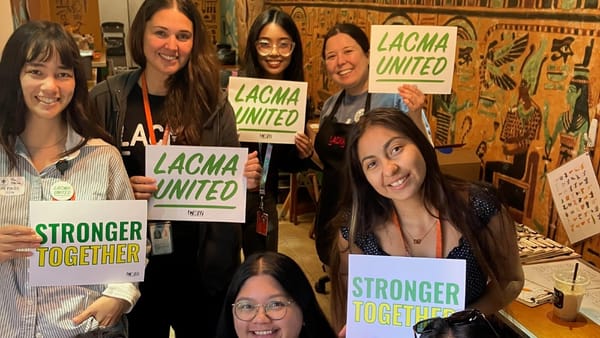Eight Los Angeles County Museum of Art employees smile and hold signs reading “LACMA United,” “Recognize Our Union,” and “Stronger Together” inside a museum workspace.