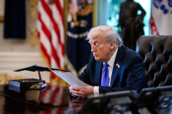 Donald Trump sits at a desk in the Oval Office, reading a document with an American flag and presidential seal visible behind him.