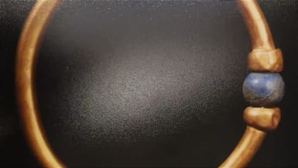 A gold ancient Egyptian bracelet with a blue bead is pictured against a black background