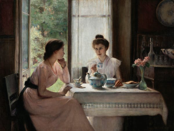 Two women sit at a sunlit table having tea beside an open doorway, with one woman leaning on her hand and listening as the other smiles and holds a teacup.