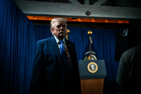 Donald Trump stands in a dimly lit room beside a podium with the presidential seal, illuminated by dramatic shadows against blue curtains.