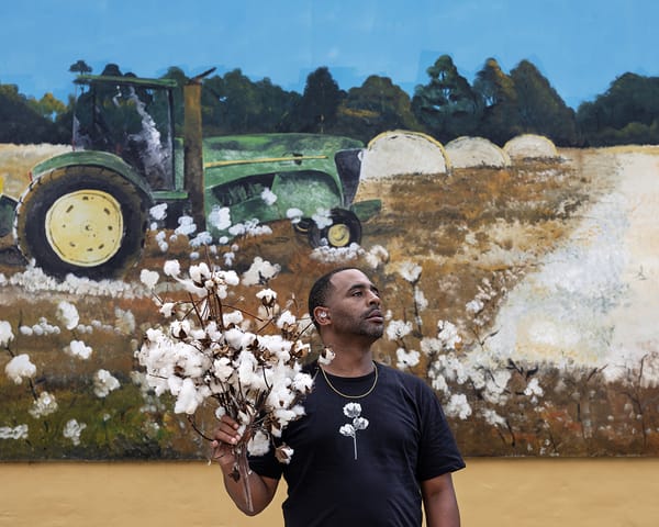 A man holds a bundle of cotton branches while standing before a mural of a cotton field and green tractor under a bright blue sky.