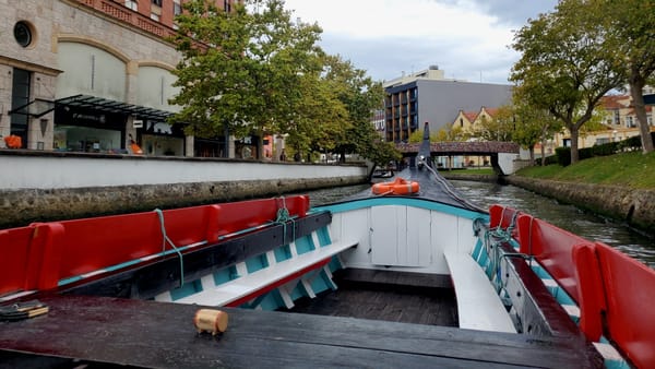 A view from inside a colorful wooden boat moving through a narrow canal lined with trees, buildings, and a small pedestrian bridge ahead.