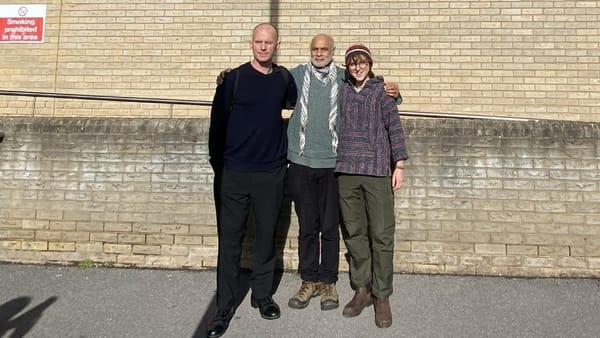 Three people stand outside against a tan brick wall in sunlight, smiling with their arms around each other.
