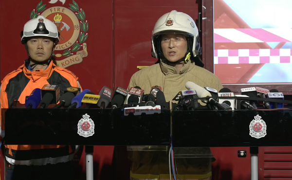 wo Hong Kong Fire Services officers stand at a podium during a nighttime press briefing, surrounded by microphones, with a fire services emblem and emergency vehicle visible behind them.