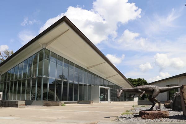 Exterior of the Museum of the Earth in Ithaca, New York, showing the modern glass-fronted building and a dinosaur sculpture outside the entrance.