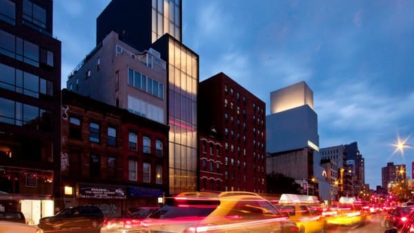 The Foster Building rises above neighboring brick structures in Manhattan at dusk. The glass-fronted gallery space, long home to Sperone Westwater, is seen amid evening traffic
