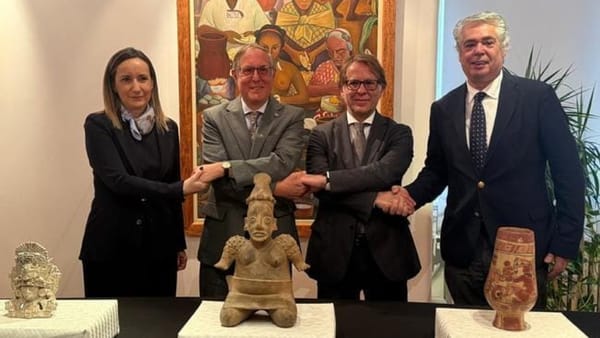 Four officials shake hands behind three pre-Hispanic artifacts displayed on tables during a restitution ceremony at the Mexican Embassy in Lisbon