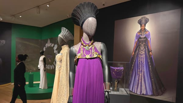 Mannequin dressed in a regal purple Wakanda gown and ornate headpiece on display in a museum gallery, with a large portrait of the character in the background and additional costumes nearby.